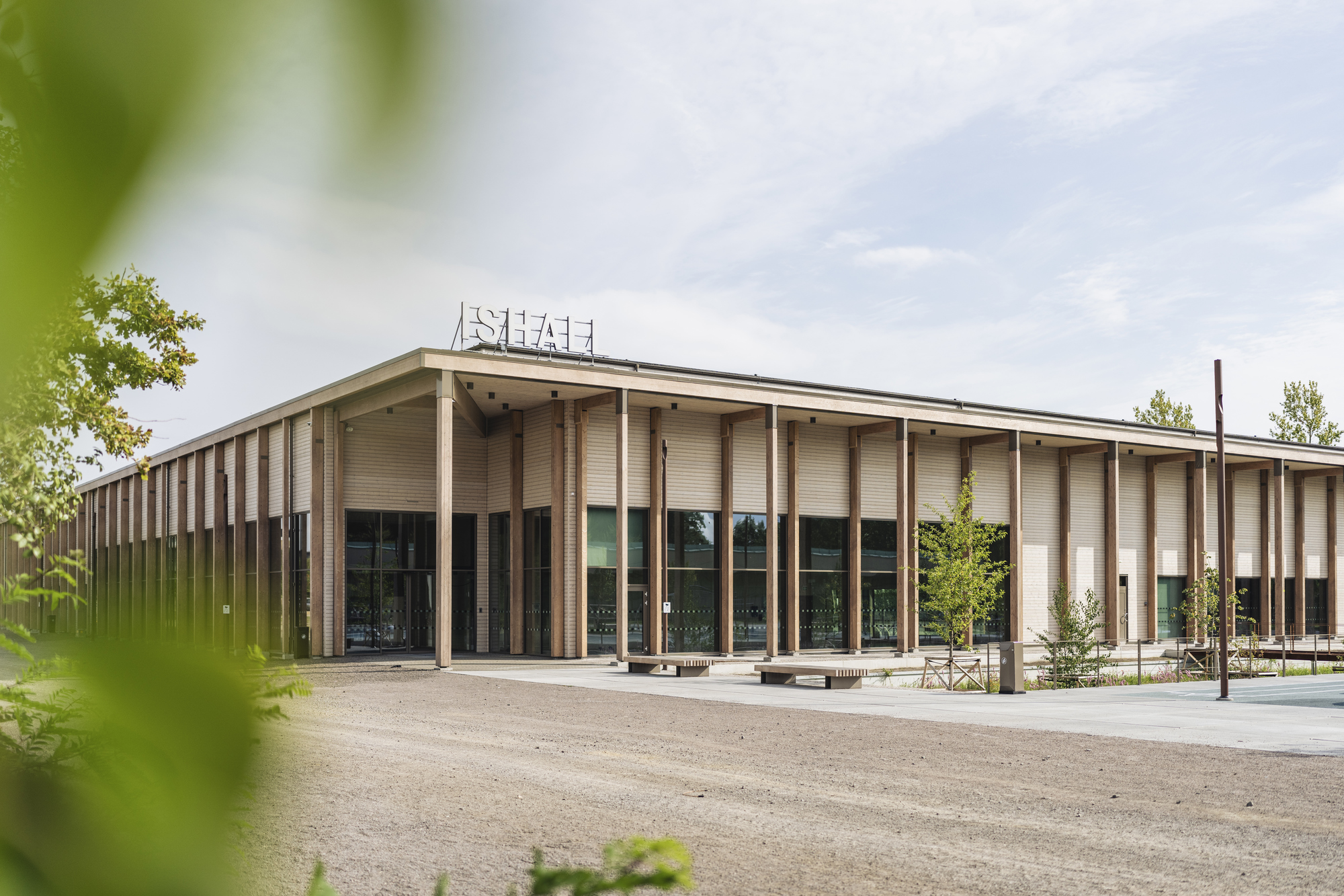En modern, ljus byggnad (ishall) med stora fönster och träpelare omgiven av träd och grönska, sedd från sidan mot en ljus himmel.