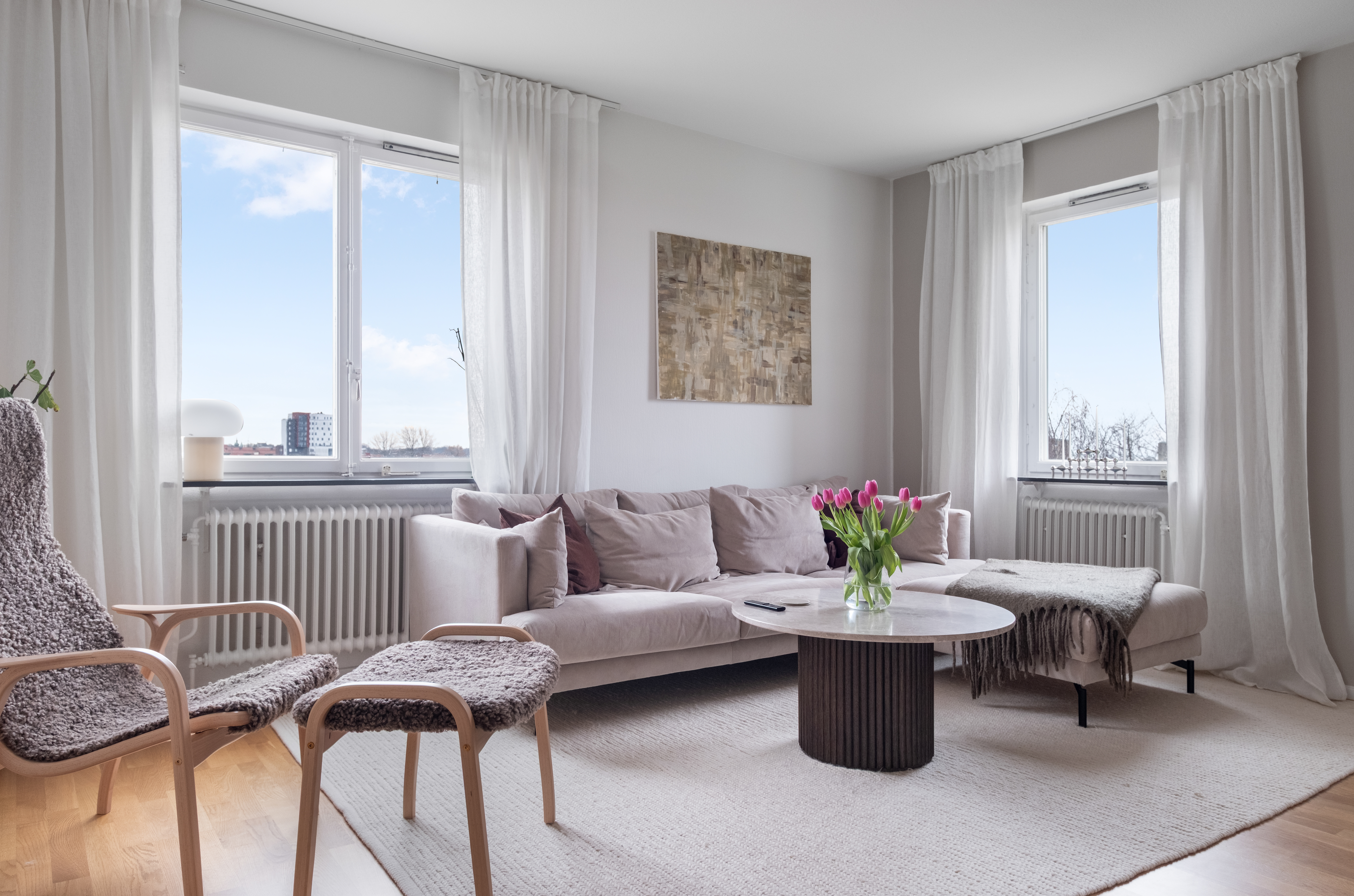 Ett ljust vardagsrum med stora fönster, vitmålade väggar och ljusa gardiner. I mitten står en ljusbeige hörnsoffa med kuddar och pläd, framför ett runt soffbord med en bukett rosa tulpaner. Till vänster syns två träfåtöljer med grå sitsar och på golvet ligger en stor, ljus matta. Utanför fönstren skymtar blå himmel och några byggnader.
