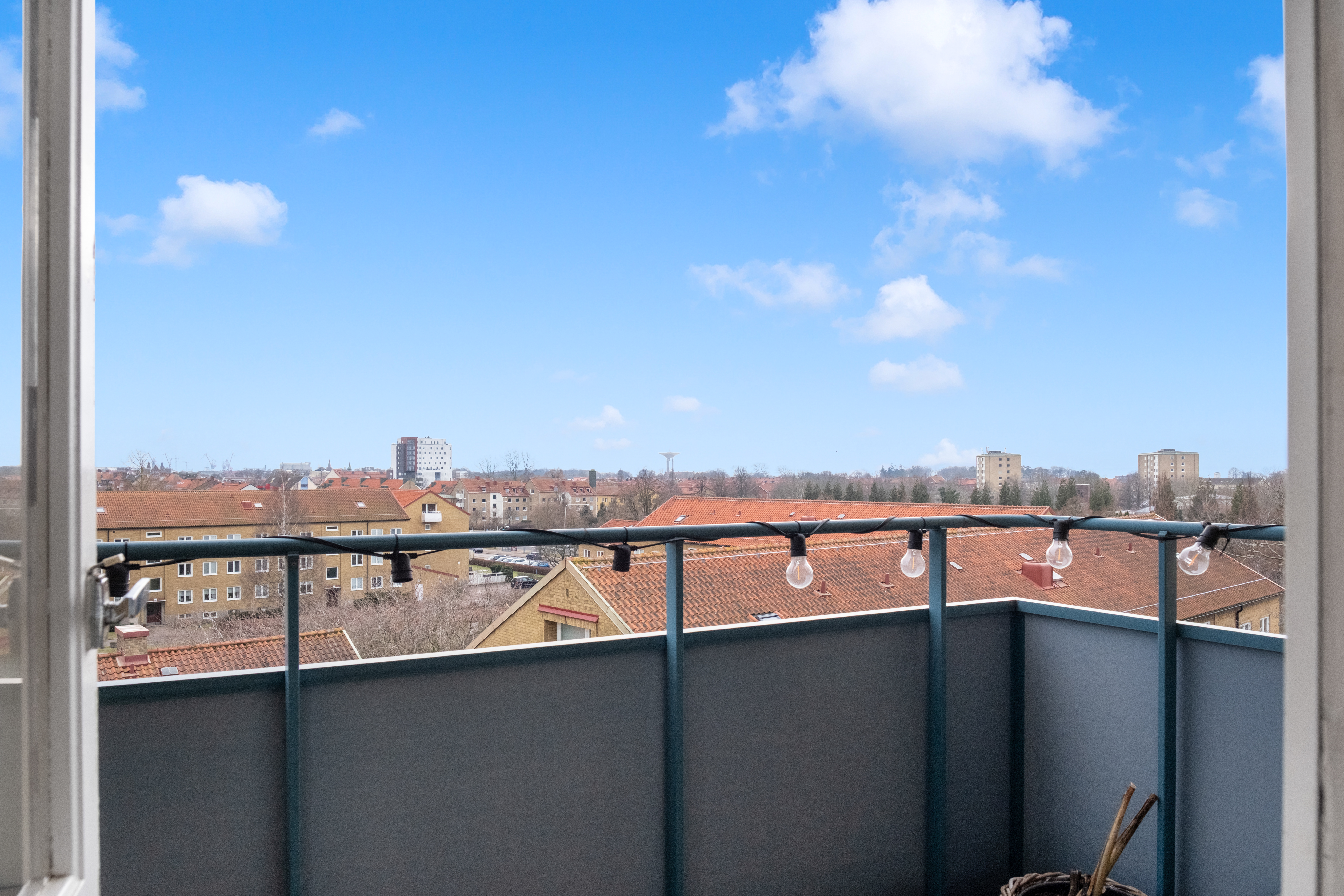 En balkong med blått räcke och ljusslinga med runda lampor, med utsikt över tak och bostadshus med röda tegeltak. I bakgrunden syns fler hus och träd under en klarblå himmel med några vita moln.