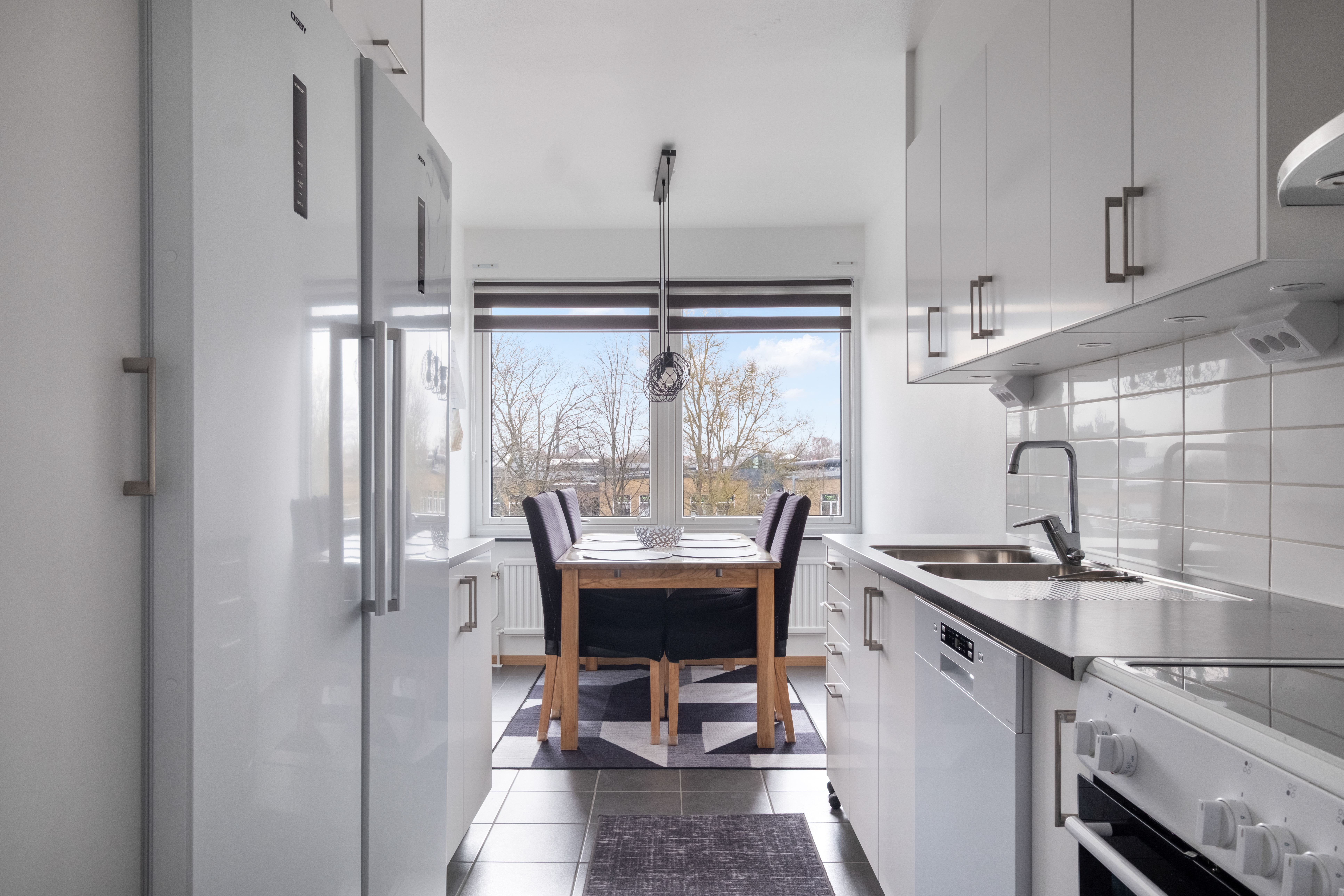 Ett ljust kök i korridorsform med vita skåp och vitvaror på båda sidor, grått klinkergolv och mörk bänkskiva. Längst in vid det stora fönstret står ett matbord i trä med fyra stoppade stolar, dukat för måltid och belyst av en taklampa, med utsikt över träd och hustak utanför.