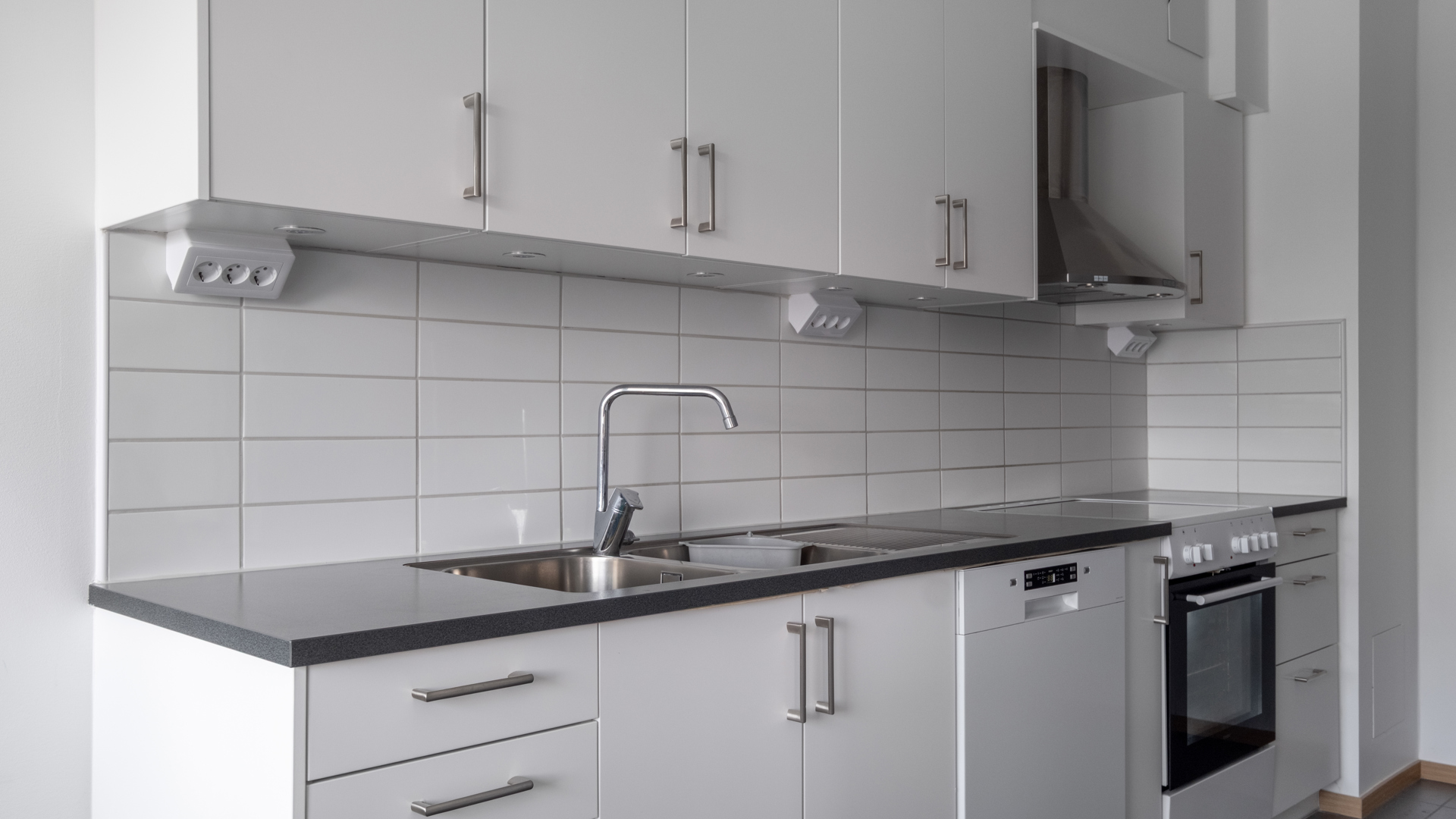 Ett modernt, ljust kök med vita släta luckor, rostfria handtag och mörk bänkskiva. Längs väggen löper en rad överskåp ovanför vit kaklad stänkskyddsvägg, och under bänken finns diskho med kromad blandare, diskmaskin och en inbyggd ugn med spishäll och rostfri fläkt.