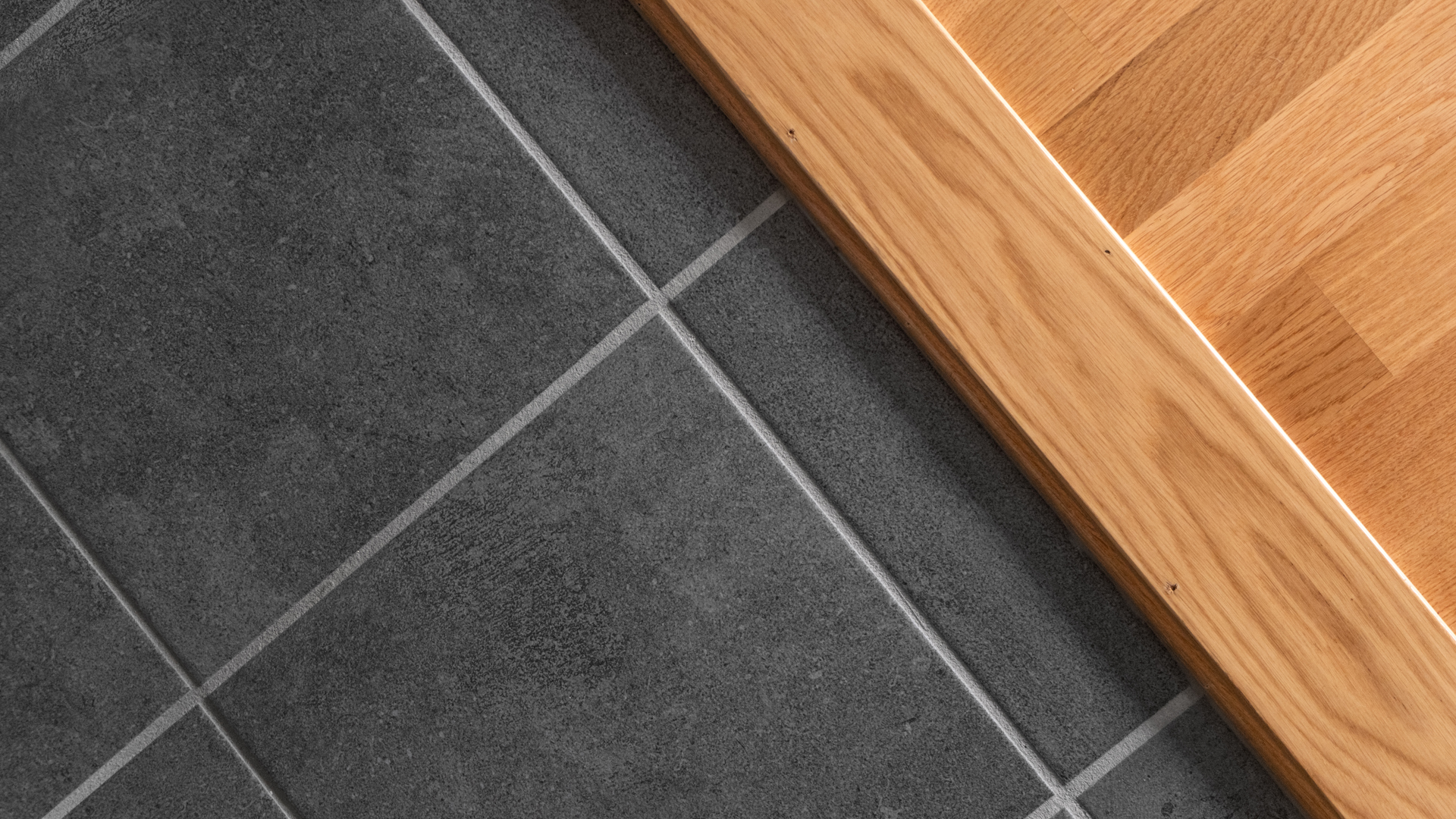 Närbild på övergång mellan mörkgrått klinkergolv med ljusa fogar och ett ljust trägolv, avskilt av en tröskel i lackat trä.