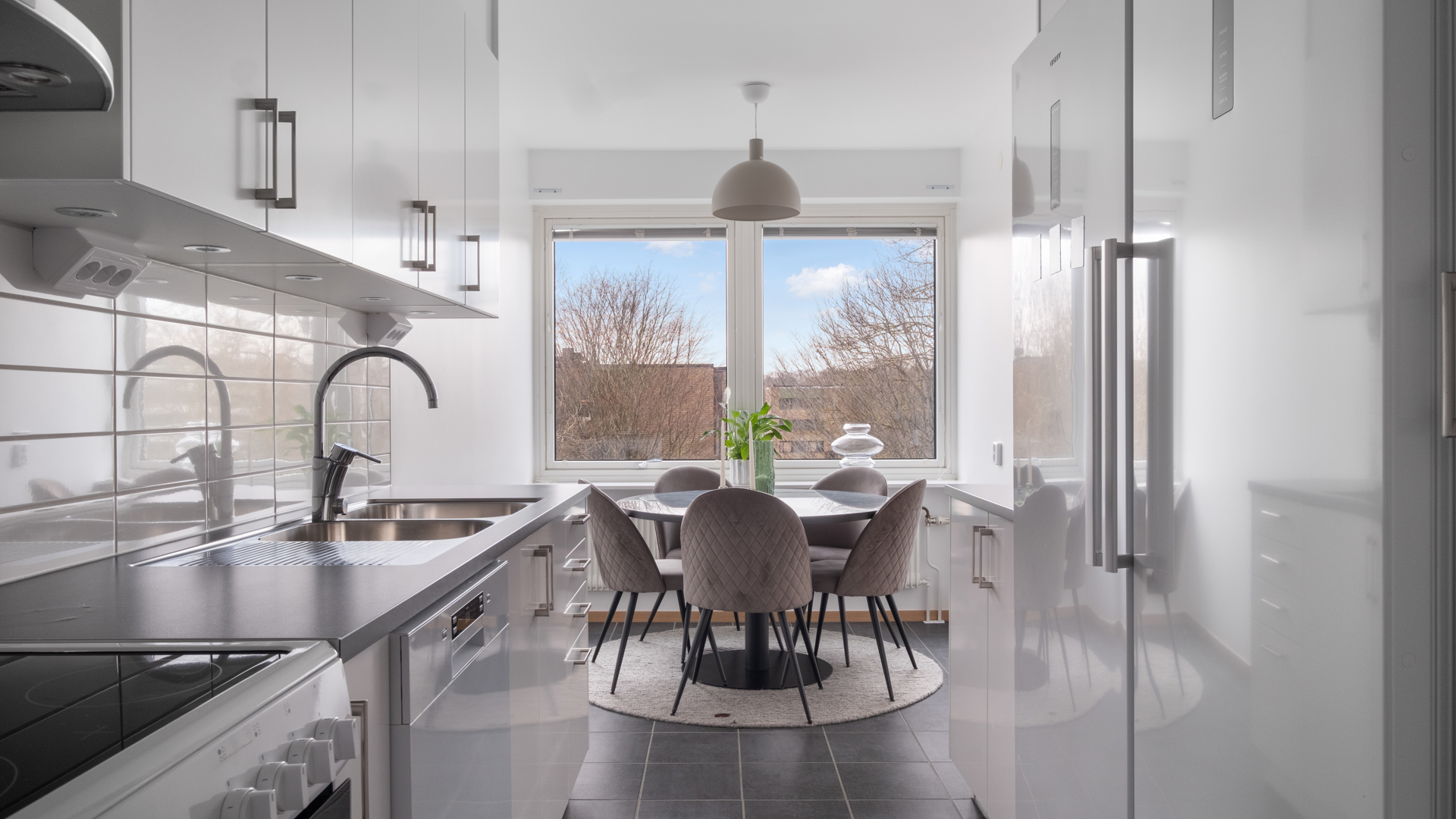 Ett ljust, avlångt kök med vita skåp och mörk bänkskiva på vänster sida, stora vita kyl- och frysskåp till höger och mörkgrått klinkergolv. Längst in vid det stora fönstret står ett runt matbord med stoppade grå stolar och gröna växter som dekoration, med utsikt mot träd och blå himmel utanför.