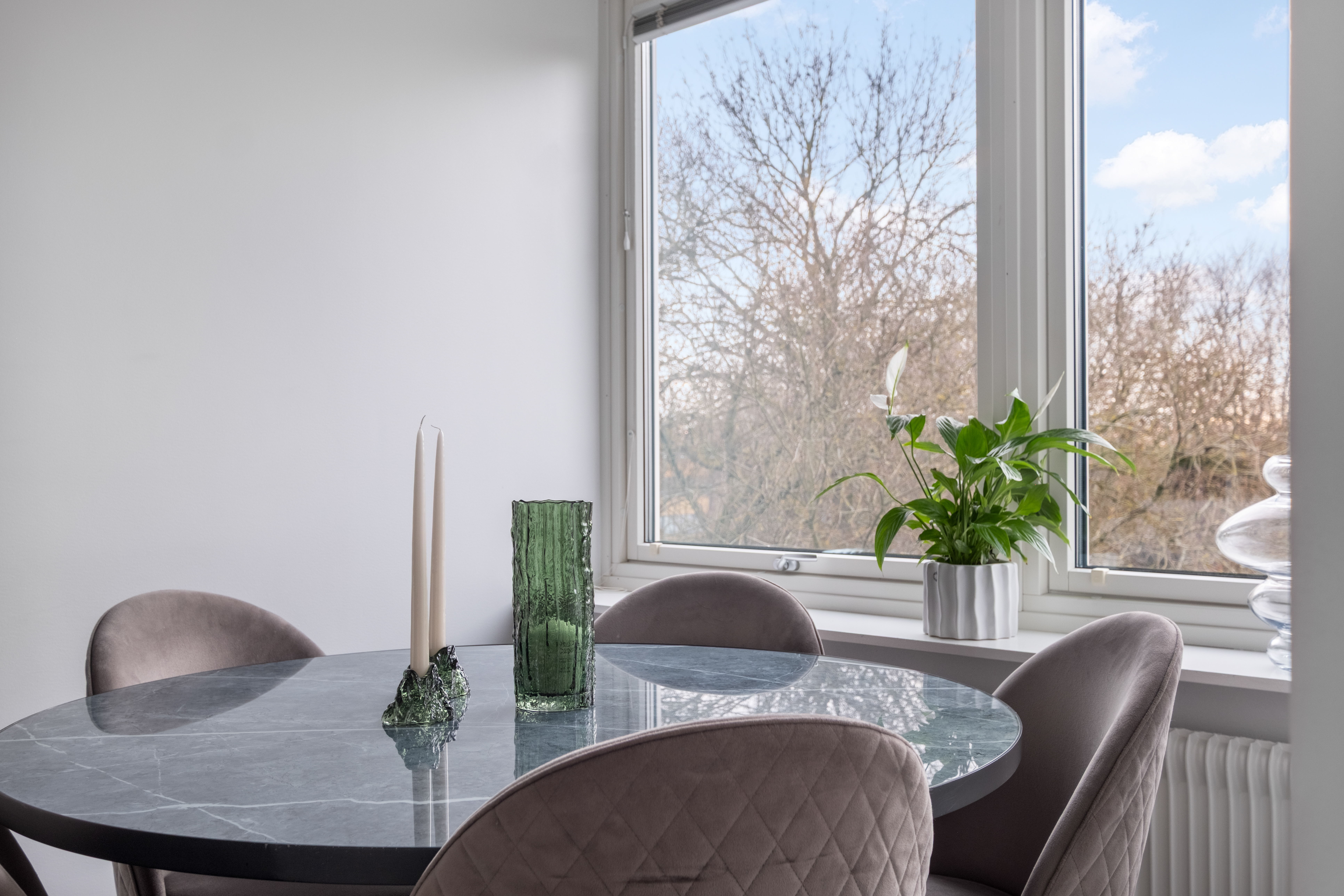 Ett närbildsfoto av en matplats med runt marmorbord omgivet av stoppade, gråbruna stolar. På bordet står två ljusa stearinljus i en grön ljusstake och en grön glasvas, och i fönstret bakom syns en grön krukväxt framför trädgrenar och blå himmel utanför.