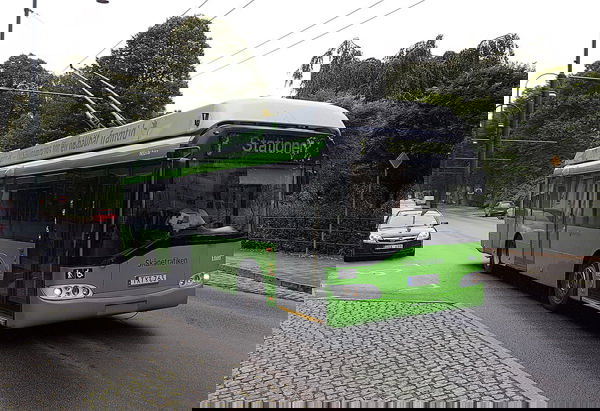 En grön trådbuss från linje 3 kör på en stadsgata, omgiven av träd och kullersten. På displayen står det "Stationen" som slutdestination. Bussen har kontaktstänger fästa i elledningar ovanför, och en bil syns bakom den i vänster körfält.