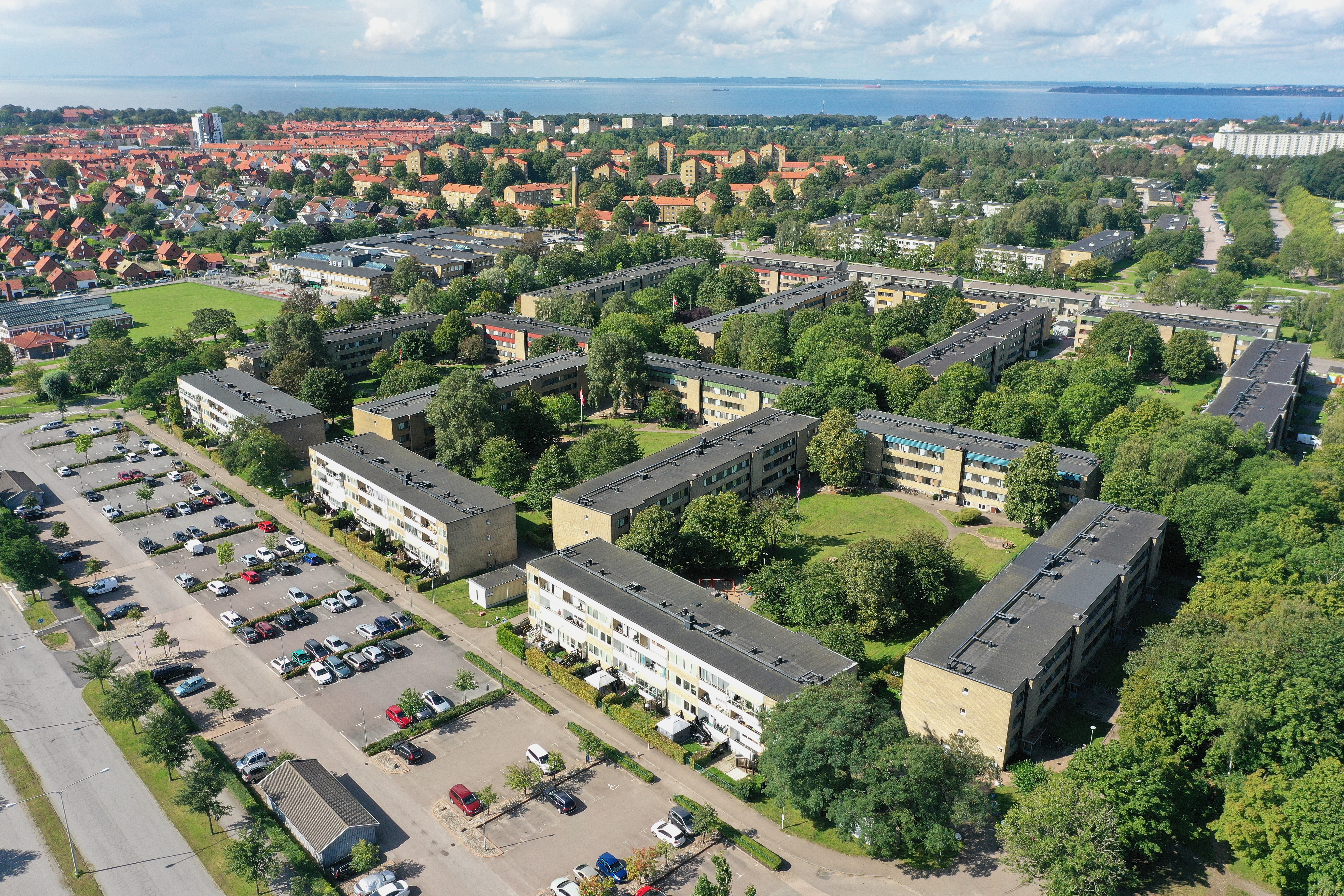 Fågelperspektiv över ett bostadsområde med flera låga, ljusgula flerfamiljshus omgivna av grönska och träd. I förgrunden syns en stor parkeringsplats med bilar. I bakgrunden breder staden ut sig med fler bostadshus, och längst bort syns havet under en blå himmel med moln.