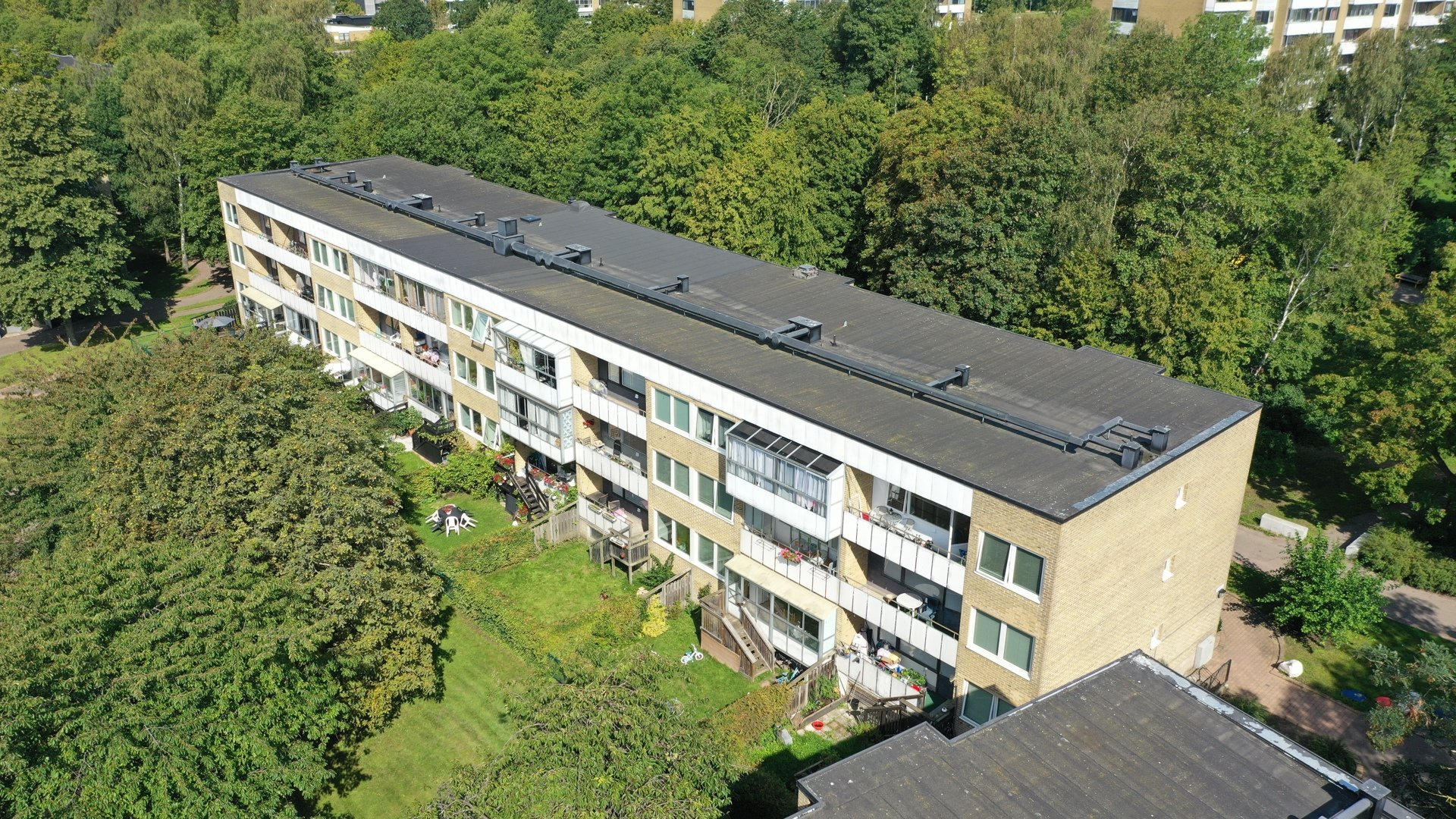Flygfoto över ett gult flerbostadshus med mörkt tak, omgivet av lummiga träd och grönska. I bakgrunden skymtar ytterligare bostadsbyggnader.