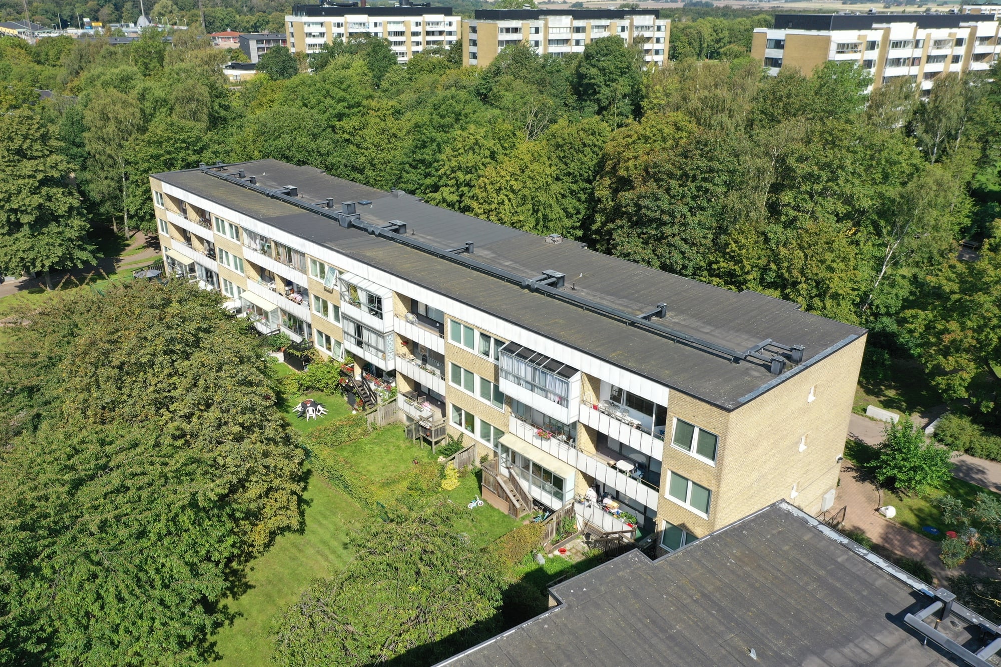 Flygfoto över ett gult flerbostadshus med mörkt tak, omgivet av lummiga träd och grönska. I bakgrunden skymtar ytterligare bostadsbyggnader.
