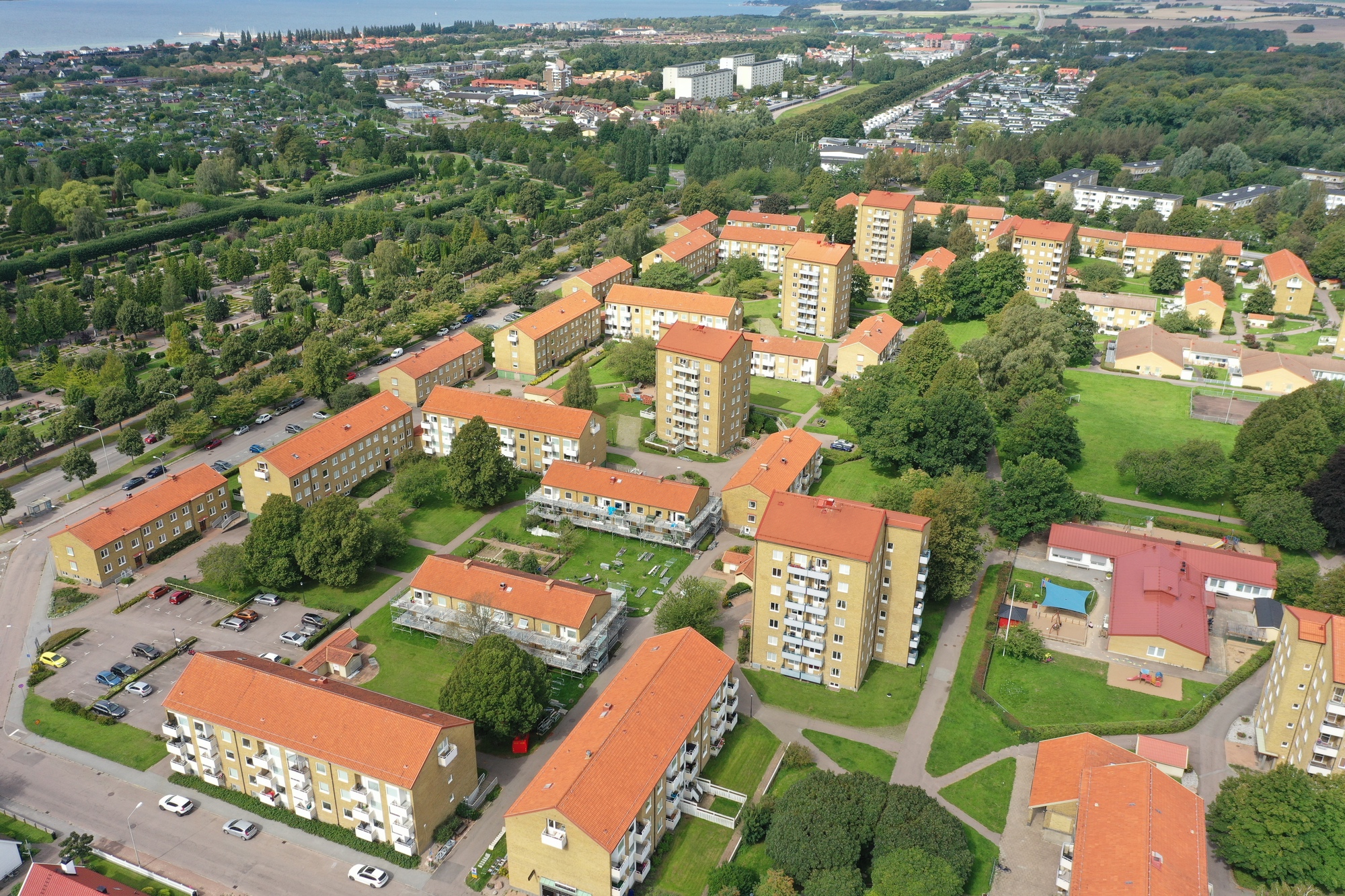 Flygfoto över ett bostadsområde med flerbostadshus, parkeringsplatser och gröna ytor. I bakgrunden syns vatten och bebyggelse.