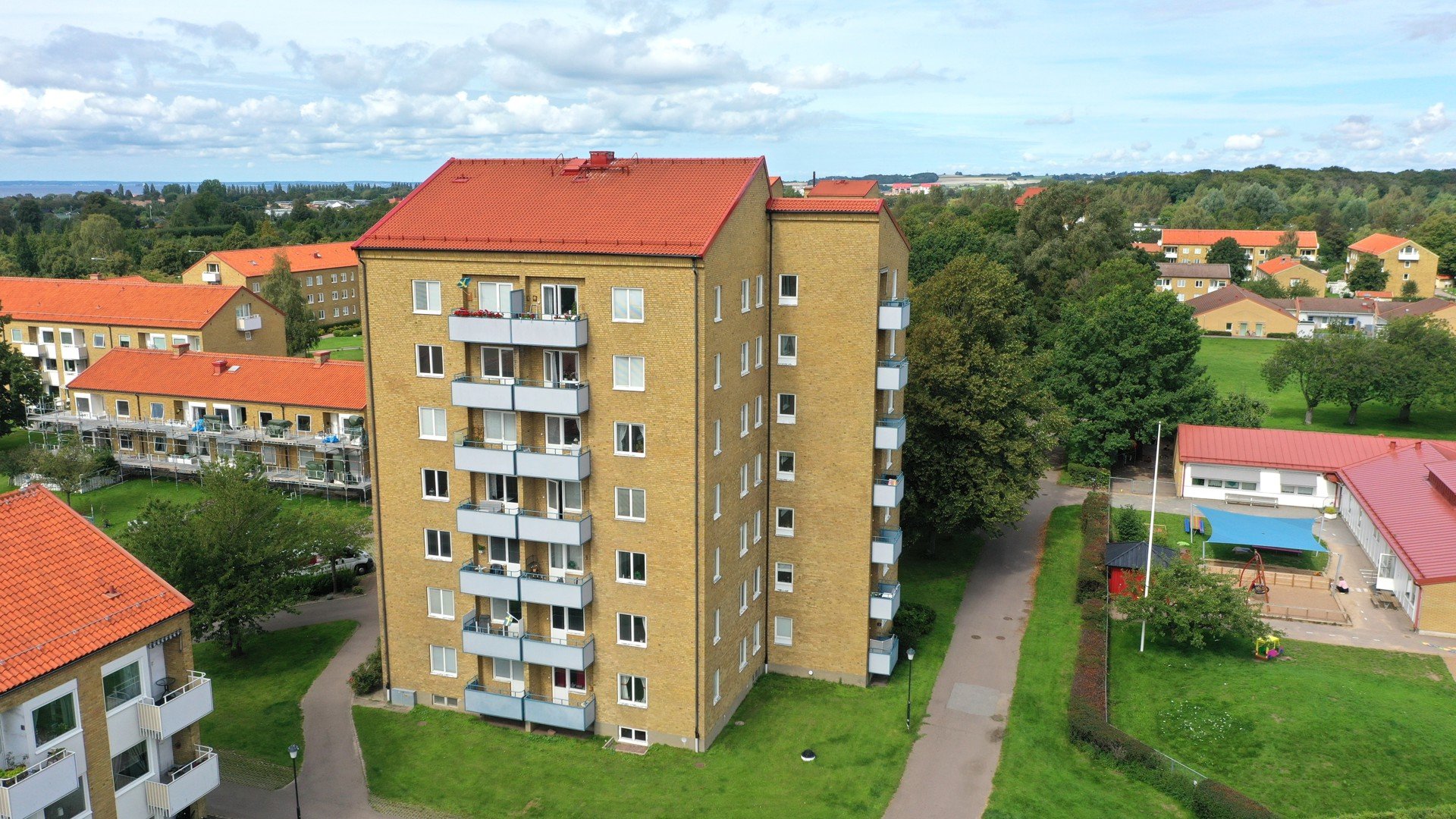 Fågelperspektiv på ett sexvånings flerfamiljshus i gult tegel med rött tegeltak. Huset har balkonger på ena sidan och omges av grön gräsmatta, gångvägar och träd. I bakgrunden syns fler bostadshus med röda tak samt ett mindre hus med pool. Himlen är delvis molnig.