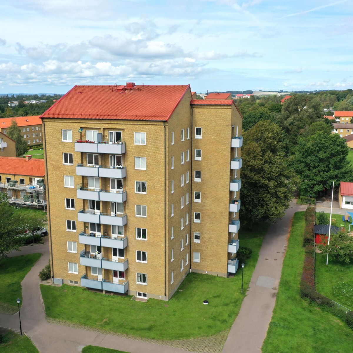 Fågelperspektiv på ett sexvånings flerfamiljshus i gult tegel med rött tegeltak. Huset har balkonger på ena sidan och omges av grön gräsmatta, gångvägar och träd. I bakgrunden syns fler bostadshus med röda tak samt ett mindre hus med pool. Himlen är delvis molnig.