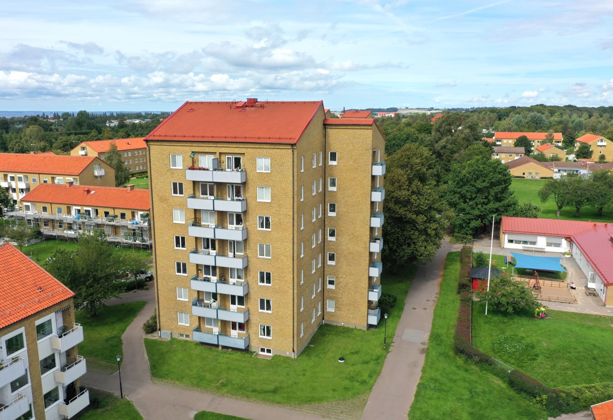 Fågelperspektiv på ett sexvånings flerfamiljshus i gult tegel med rött tegeltak. Huset har balkonger på ena sidan och omges av grön gräsmatta, gångvägar och träd. I bakgrunden syns fler bostadshus med röda tak samt ett mindre hus med pool. Himlen är delvis molnig.