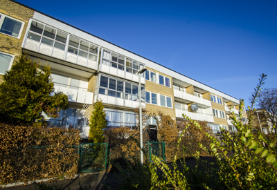 Ett trevånings flerbostadshus i gult tegel med stora inglasade balkonger. Framför huset växer välskötta buskar och träd, och en blå himmel breder ut sig ovanför byggnaden.