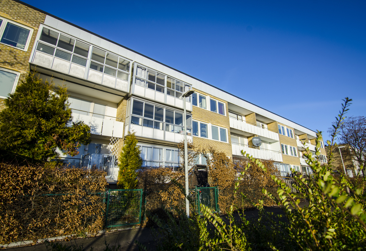 Ett trevånings flerbostadshus i gult tegel med stora inglasade balkonger. Framför huset växer välskötta buskar och träd, och en blå himmel breder ut sig ovanför byggnaden.