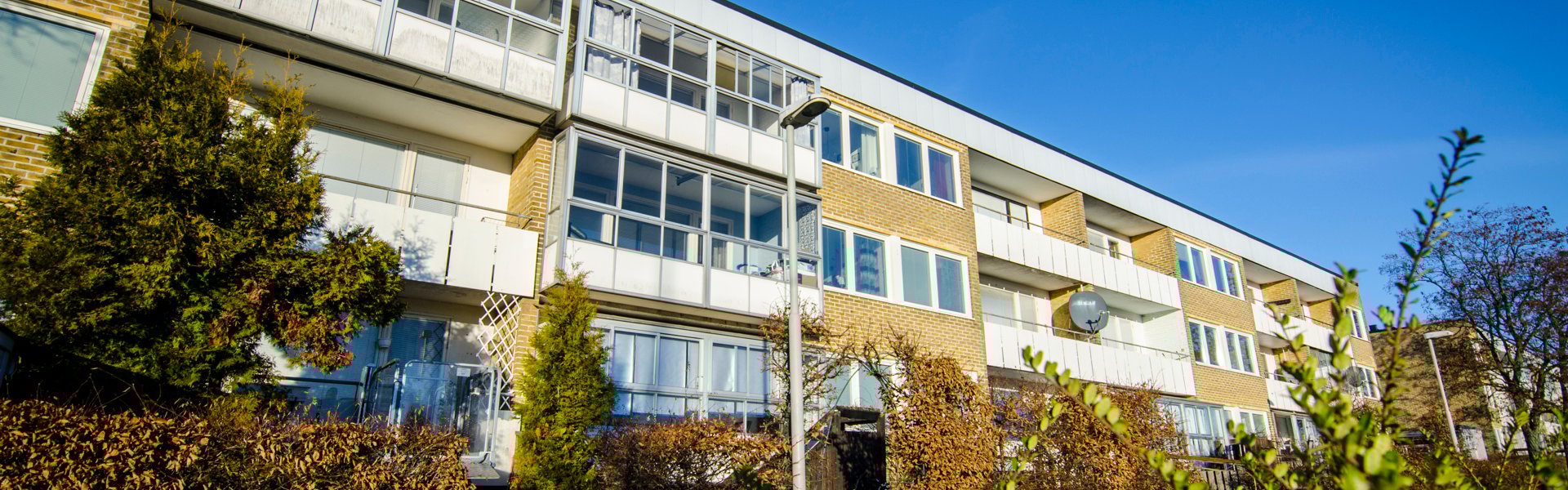 Ett trevånings flerbostadshus i gult tegel med stora inglasade balkonger. Framför huset växer välskötta buskar och träd, och en blå himmel breder ut sig ovanför byggnaden.