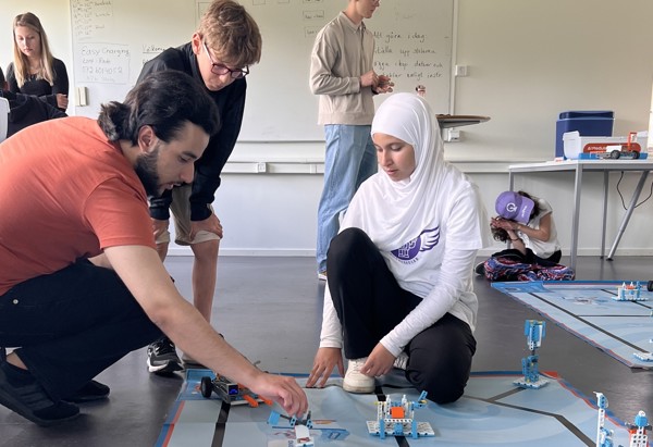 En grupp unga personer arbetar tillsammans med robotik och tekniska byggsatser på golvet i ett klassrum, omgivna av datorer och utrustning.