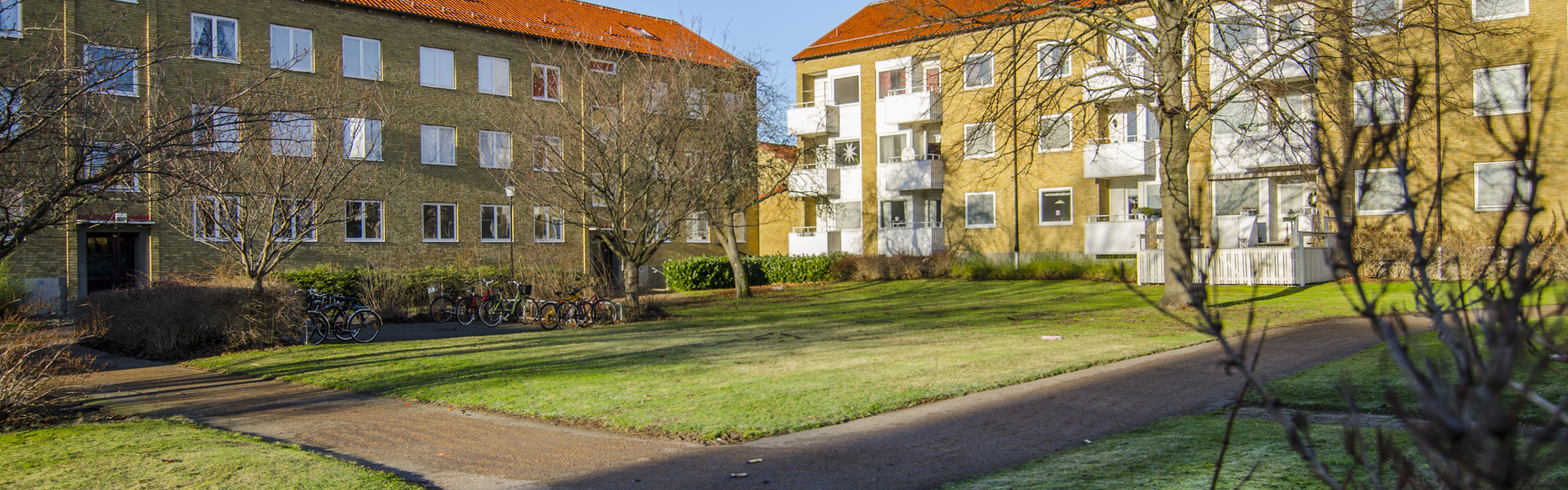 Två tegelklädda flerfamiljshus med röda tak omger en grön innergård. Det är en solig dag med frost på marken och några träd utan löv, vilket tyder på tidig vår eller sen höst.