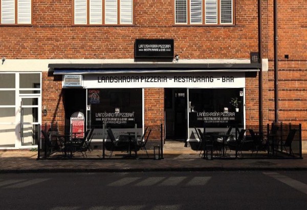 En restaurang i bottenvåningen av en tegelbyggnad med uteservering. Utanför står flera bord och stolar uppställda längs trottoaren. Solen skiner och det är tomt på gäster just nu.