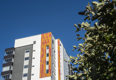 Ett modernt flerfamiljshus med färgglada vertikala paneler i orange och gult, mot en klarblå himmel. En del av en grön buske syns i förgrunden till höger i bilden.