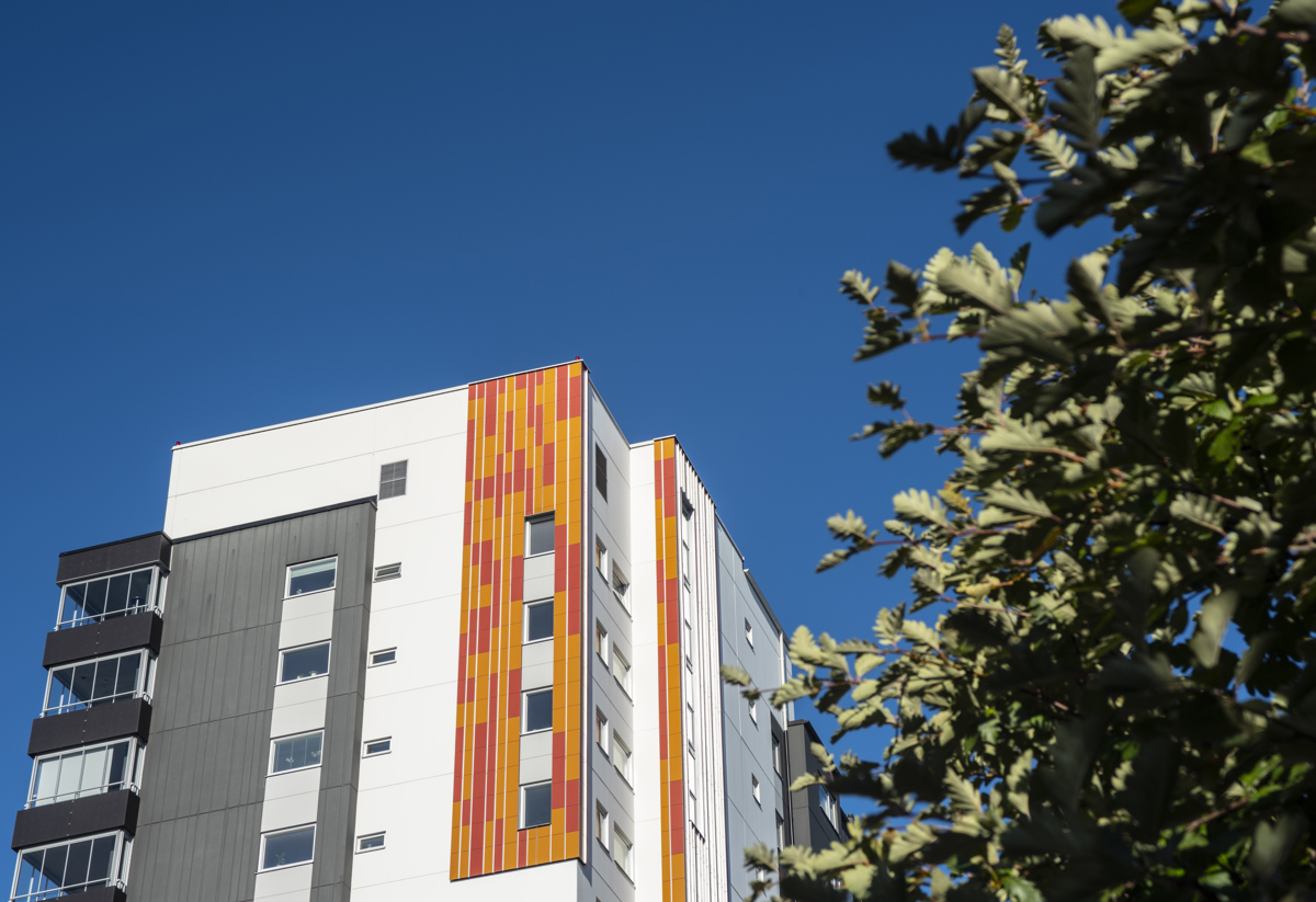 Ett modernt flerfamiljshus med färgglada vertikala paneler i orange och gult, mot en klarblå himmel. En del av en grön buske syns i förgrunden till höger i bilden.