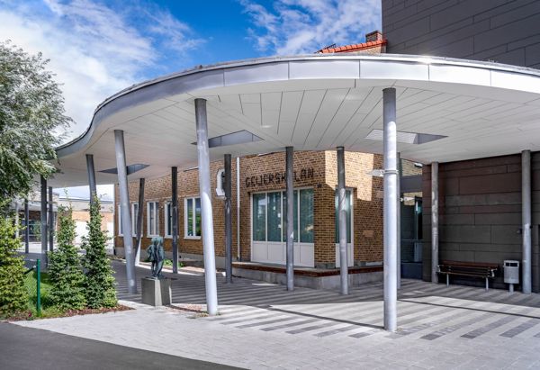 Entrén till en modern byggnad med ett böjt tak som vilar på smala pelare. Fasaden är av tegel. Området är stenbelagt och omges av träd och buskar. Himlen är delvis molnig med blå himmel i bakgrunden.