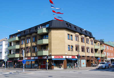 Ett gathörn med ett fyravåningshus i gult tegel, med balkonger och butikslokaler i bottenplan. Röda, vita och blå vimplar svävar i luften mot en klarblå himmel.