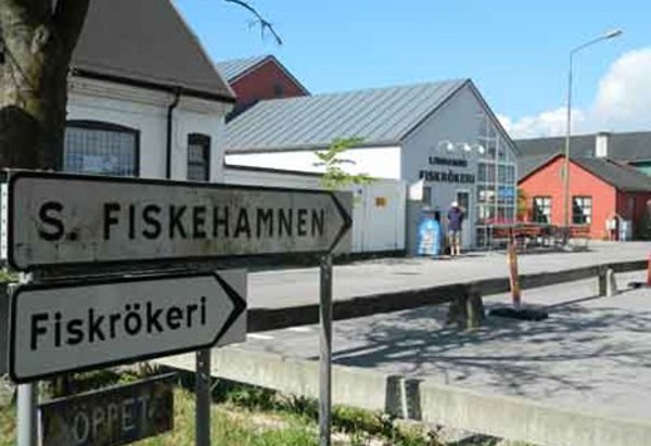 Skyltar som visar vägen till "Södra fiskehamnen" och ett "Fiskrökeri" i förgrunden. I bakgrunden syns en vit byggnad med glasfasad där det står "Limhamns Fiskrökeri", omgiven av fler låga byggnader i olika färger. Några personer syns utanför byggnaden, och himlen är klar med några moln.