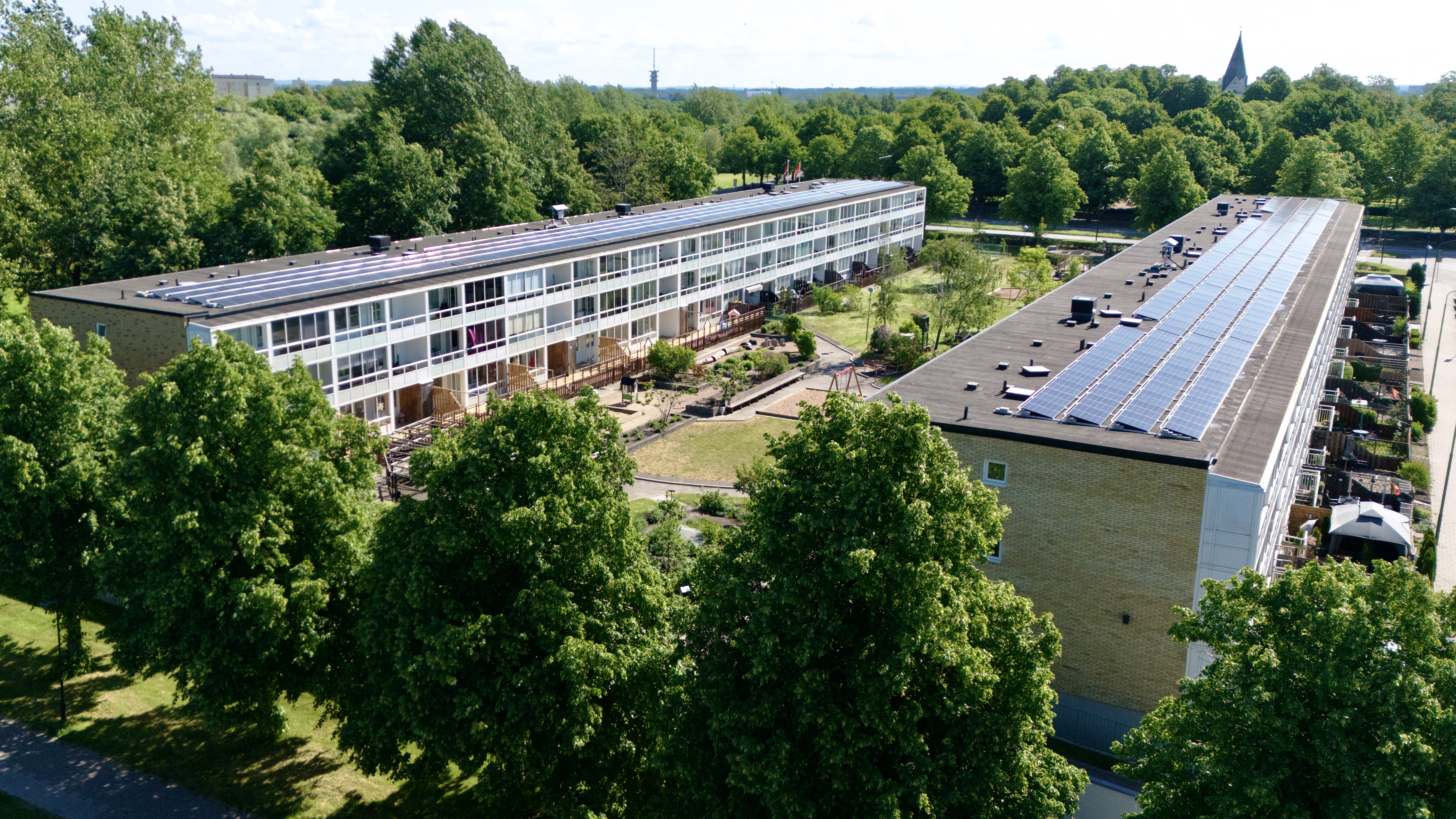 Flygbild över låga flerbostadshus med solpaneler på taken, omgivna av grönskande träd och gårdsmiljö.