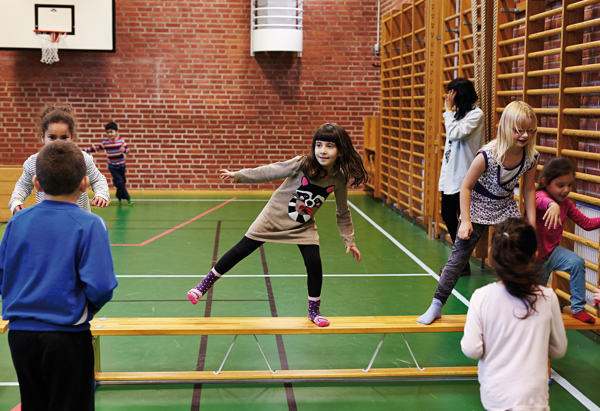 Ett gäng barn leker och tränar balans i en gymnastiksal. Ett barn går balanserande på en plint, medan andra barn tittar på eller väntar på sin tur. Gymnastiksalen har grönt golv, röda tegelväggar och ribbstolar längs väggarna.