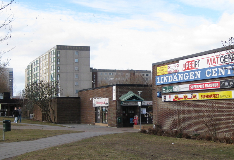 Lindängen centrum med skyltar som visar olika butiker och restauranger. I bakgrunden syns höghus med flera våningar. Omgivningen har gångvägar, gräsytor och träd utan löv, vilket tyder på att bilden är tagen under höst eller tidig vår.