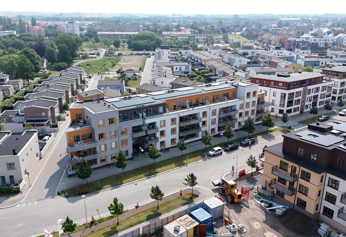 Flygbild över ett nybyggt bostadsområde med flerbostadshus, radhus, gator och grönska.