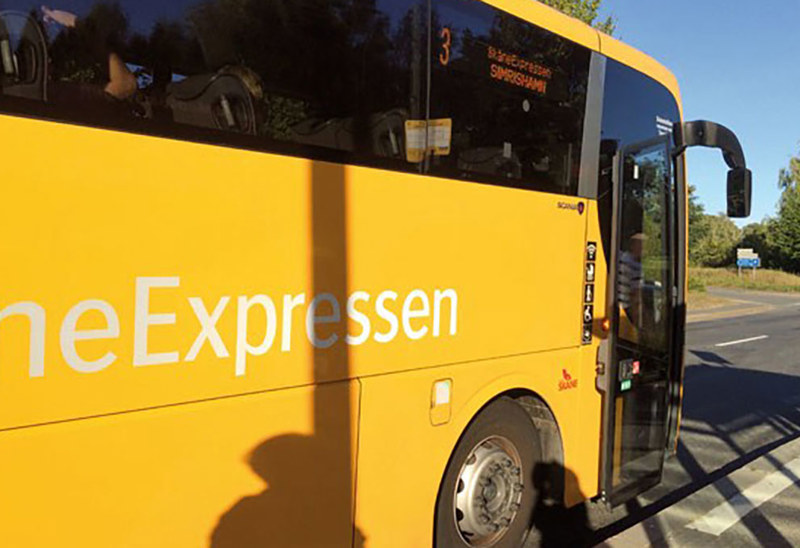 En gul buss från SkåneExpressen står stilla vid en hållplats en solig dag. Bussens dörr är öppen och en skugga från en person syns på sidan av bussen. Destinationen på skylten visar 'Simrishamn'.