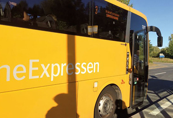 En gul buss från SkåneExpressen står stilla vid en hållplats en solig dag. Bussens dörr är öppen och en skugga från en person syns på sidan av bussen. Destinationen på skylten visar 'Simrishamn'.