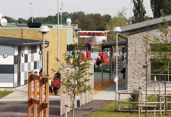 Ett modernt skolområde med flera byggnader i olika stilar – en rutmönstrad fasad, en i gult tegel och en i ljusgrått tegel. På gården syns barn som leker vid ett färgglatt lekområde. Mellan byggnaderna går en gångväg kantad av unga träd och lampstolpar. I bakgrunden syns grönska och ett staket som omger området.