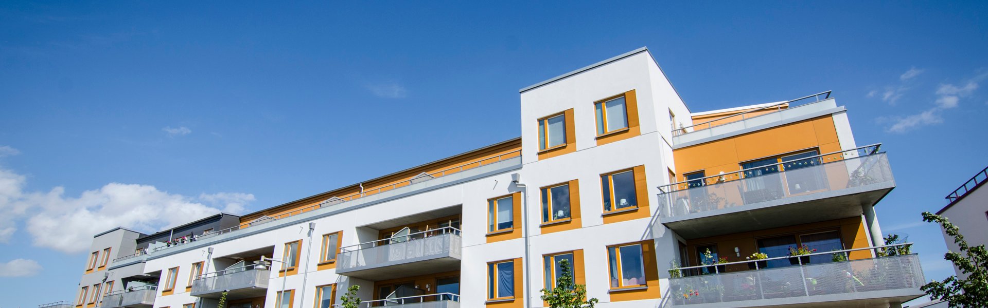 Ett modernt flerfamiljshus i vitt och orange med flera balkonger och stora fönster. Huset har fyra våningar och står längs en asfalterad gata med parkerade bilar och unga träd planterade på trottoaren. Himlen är klarblå med några moln.