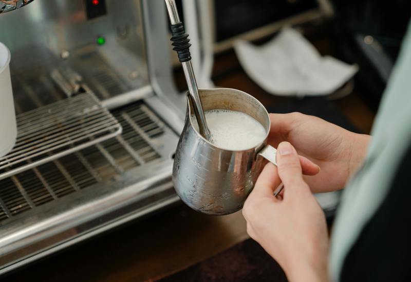 En person skummar mjölk med hjälp av en ångstav från en espressomaskin. Personen håller i en metallkanna med varm mjölk och ångan syns tydligt.