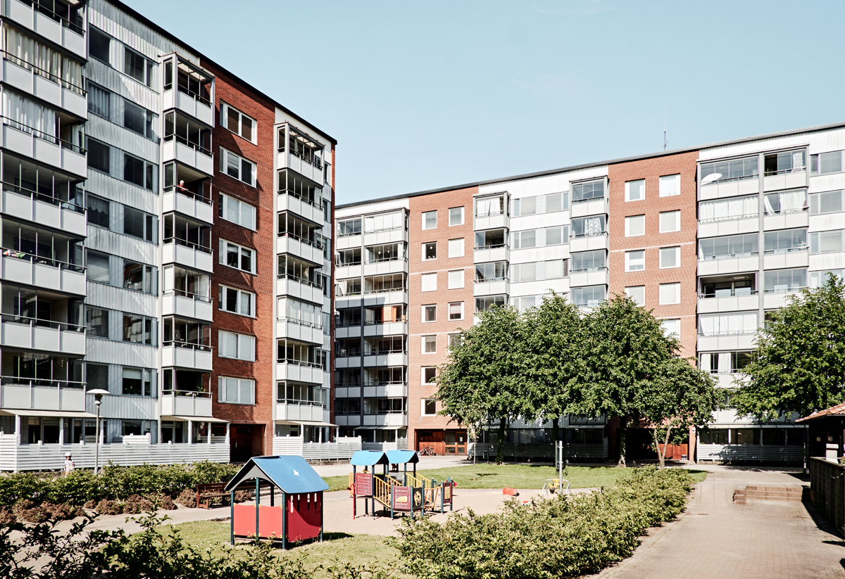 Innergård mellan höga flerfamiljshus med röd och vit fasad. På gården finns en liten lekplats med färgglada lekstugor, en stor grön träd och gångvägar som leder genom området. Det är en solig dag med klarblå himmel.