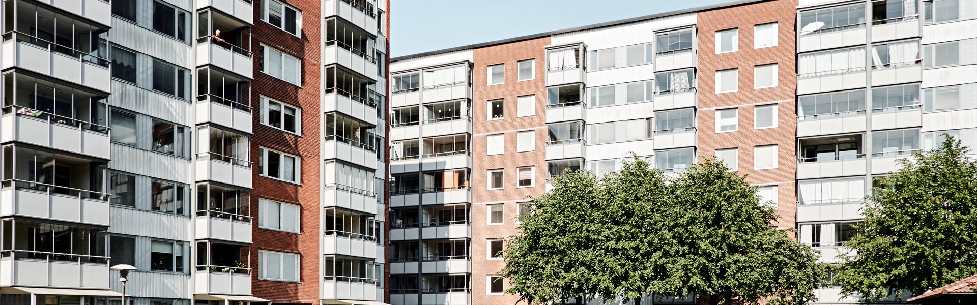 Innergård mellan höga flerfamiljshus med röd och vit fasad. På gården finns en liten lekplats med färgglada lekstugor, en stor grön träd och gångvägar som leder genom området. Det är en solig dag med klarblå himmel.
