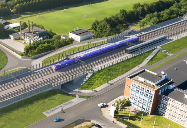 Ett pågatåg passerar genom ett grönområde nära en stad. Tåget kör på en upphöjd järnväg bredvid ett modernt kontorshus. I bakgrunden syns fotbollsplaner, flerbostadshus och mycket grönska.
