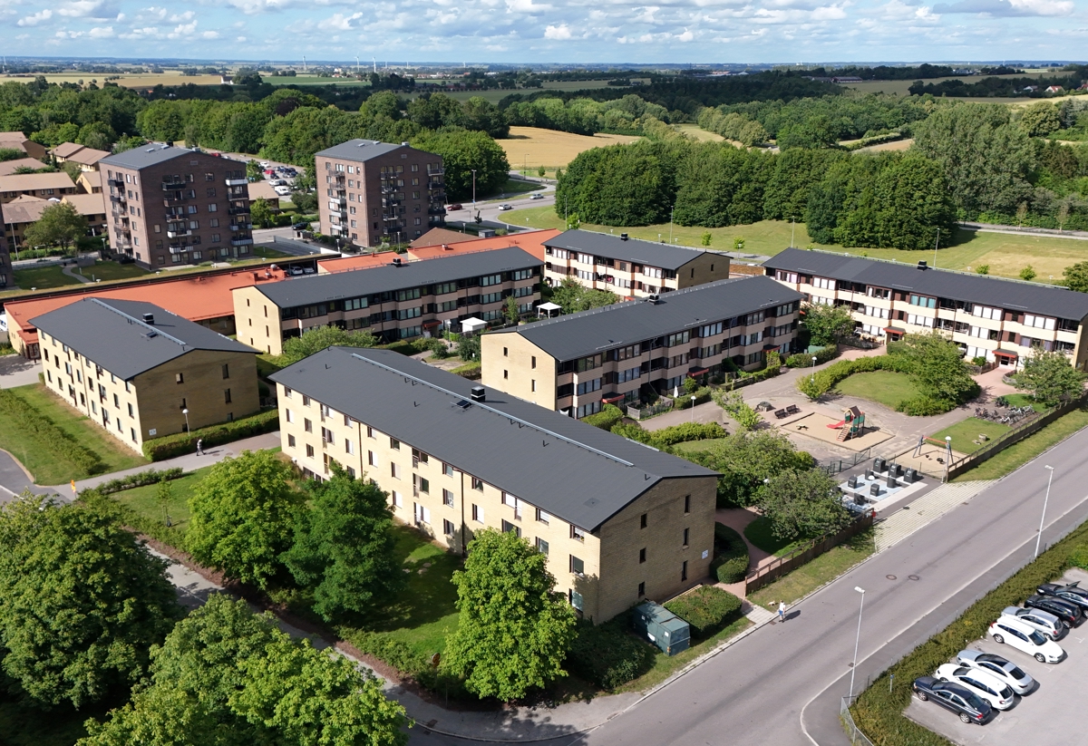 Flygbild över ett bostadsområde med låga flerbostadshus, innergårdar, lekplatser, grönska och öppna landskap i bakgrunden.