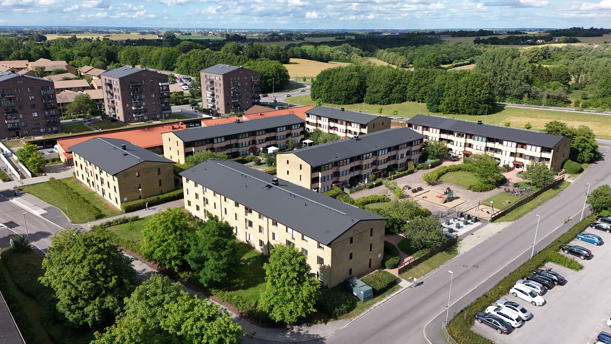 Flygbild över ett bostadsområde med flera flerbostadshus, innergårdar, parkeringar och omgivande grönområden. I bakgrunden syns åkrar, skog och en väg under en delvis molnig himmel.