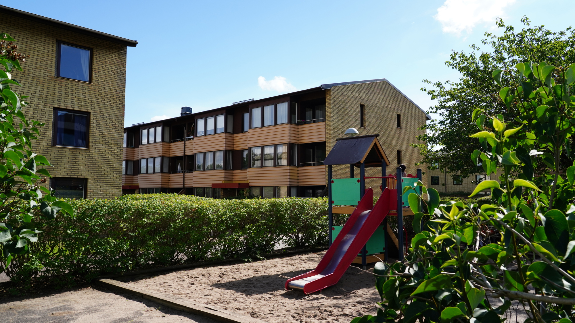 En lekplats med rutschkana och sandlåda framför flerbostadshus i gult tegel och träfasad. Området omges av häckar och grönska under en solig himmel.