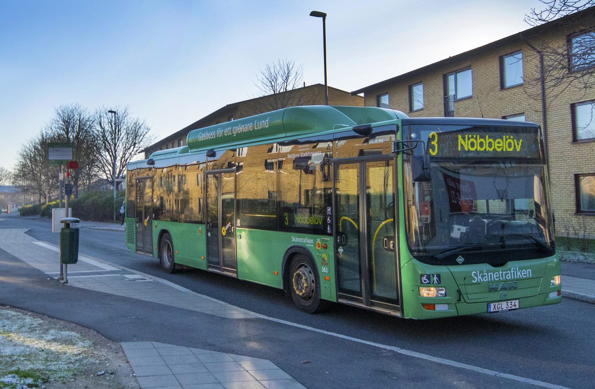 En grön stadsbuss från Skånetrafiken står vid en hållplats. Bussen är på linje 3 med destination "Nöbbelöv", enligt skylten framtill. I bakgrunden syns ett flerfamiljshus i gul tegel, och träden runtomkring är kala, vilket antyder att det är vinter eller sen höst. Det är tidig morgon eller sen eftermiddag, med ett kallt blått ljus och lite frost på marken.