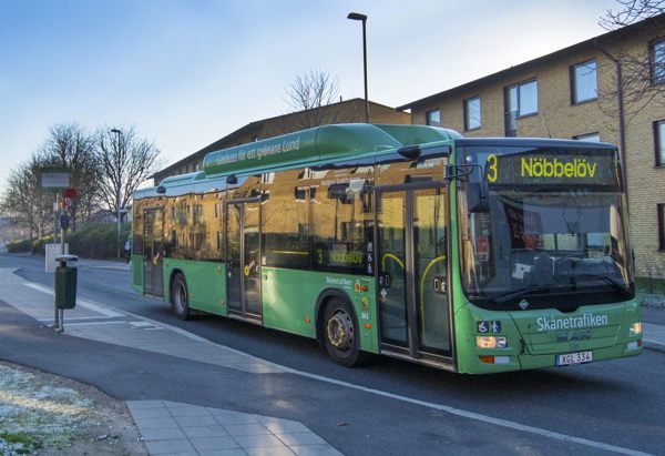En grön stadsbuss från Skånetrafiken står vid en hållplats. Bussen är på linje 3 med destination "Nöbbelöv", enligt skylten framtill. I bakgrunden syns ett flerfamiljshus i gul tegel, och träden runtomkring är kala, vilket antyder att det är vinter eller sen höst. Det är tidig morgon eller sen eftermiddag, med ett kallt blått ljus och lite frost på marken.