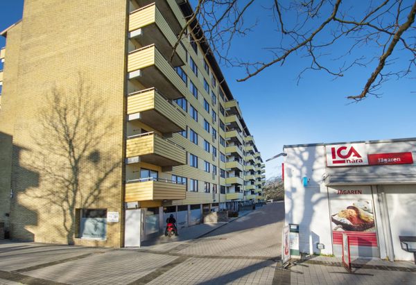 Ett höghus i gul tegel med flera balkonger längs fasaden. Till höger i bilden syns en liten butik med skylten "ICA nära". Det är en solig dag med blå himmel, och kala trädgrenar sträcker sig in i bilden från höger. Stenar täcker marken framför byggnaderna.