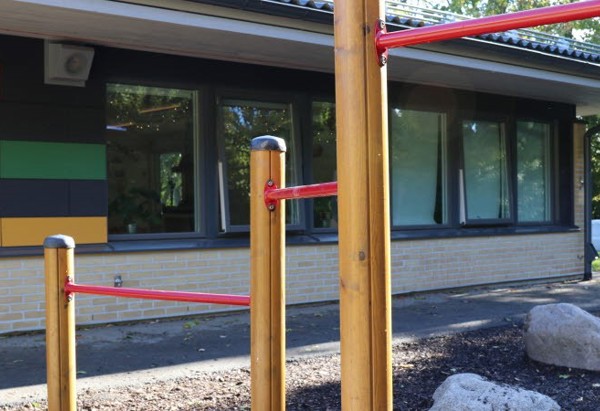 Ett lekredskap med röda metallstänger och trästolpar framför en byggnad med fönster och färgglad fasad. Byggnaden har ljus tegelfasad med dekorativa paneler i olika färger som rött, blått, grönt och svart. I förgrunden finns stora stenar och barkflis på marken. Träd syns i bakgrunden.