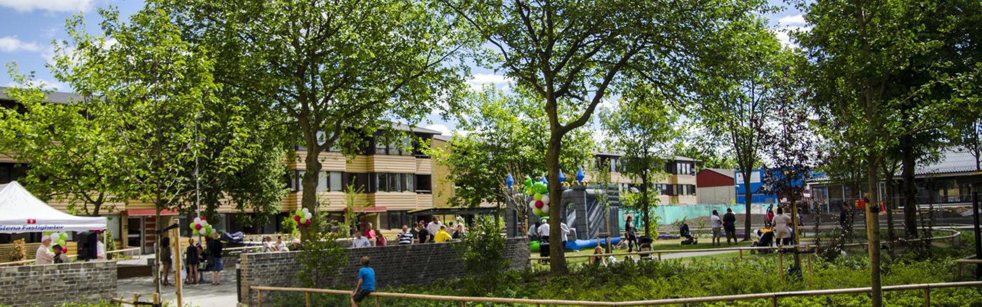Grönskande parkmiljö med asfalterade gångvägar, träd och buskar. I bakgrunden syns lekplats, människor som vistas i området samt flerbostadshus. Soligt väder med blå himmel och vita moln.