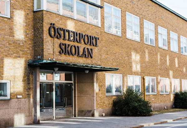 En skolbyggnad i gult tegel med skylten "Österportskolan" ovanför huvudentrén. Byggnaden har stora fönster i två våningar och ett grönt takutsprång över entrédörrarna. Solens reflexer syns på fasaden och några buskar växer längs väggen vid trottoaren.