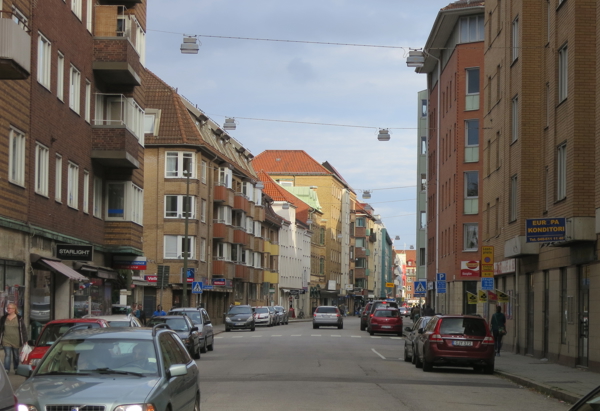 En stadsgata kantad av flerbostadshus i olika färger och material, med fasader i rött, gult och beige tegel. Flera bilar är parkerade längs gatan och några är i rörelse. Gatuscenen ramas in av trottoarer med butiker och skyltar. Gatubelysning hänger i luften mellan byggnaderna, och himlen är delvis molnig.
