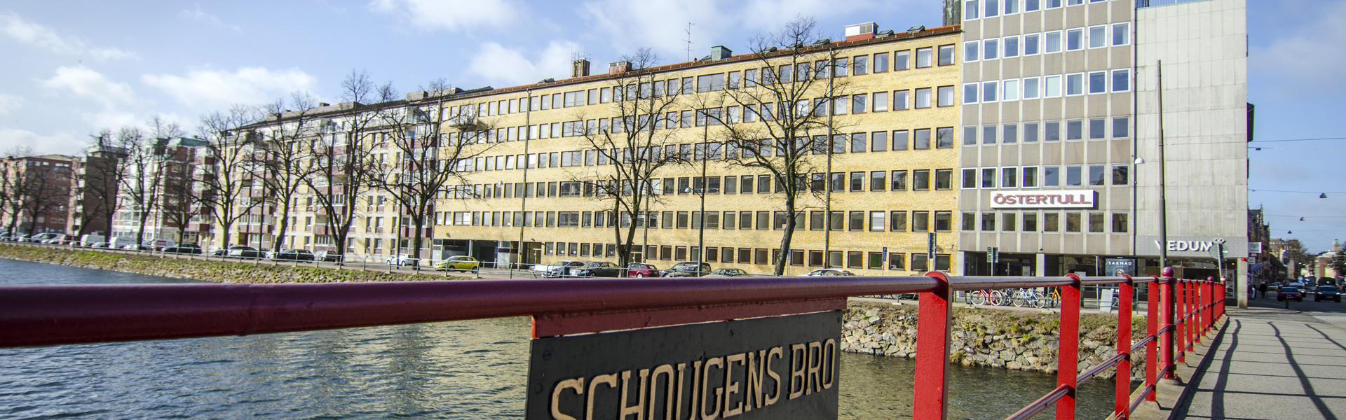En vy från en bro med röd räcke, där en skylt med texten "Schougens bro 1949" syns i förgrunden. I bakgrunden ser man en lång kontors- eller bostadsbyggnad med många fönster, belägen vid en kanal eller ett vattendrag. Solen skiner och himlen är delvis molnig.